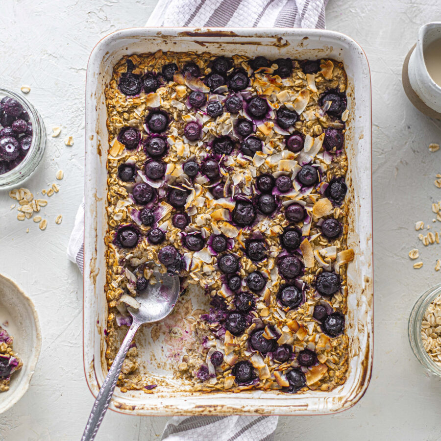 A baking dish of baked oatmeal topped with blueberries and oats, with a spoon—plant-based cheesecake style. Nearby: porridge oats, blueberries, oats, and a jug of plant-based milk.