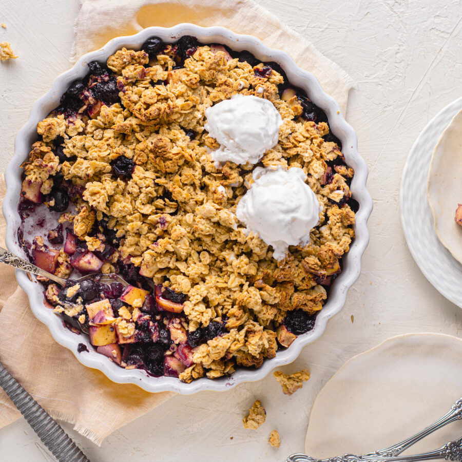 Round dish of Apple Blueberry Crumble with oat topping and two scoops of plant-based vanilla ice cream, plates, and cutlery nearby.