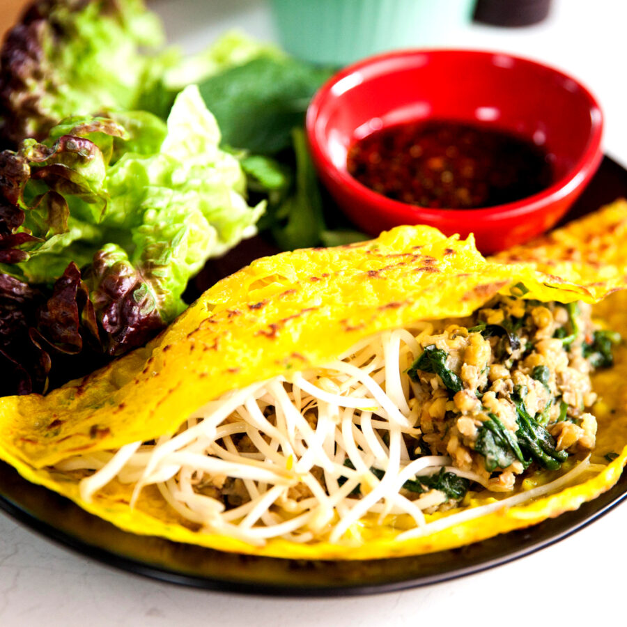 A Vietnamese Bánh Xèo turmeric pancake filled with mung beans, bean sprouts, herbs, and plant-based protein, served with lettuce and a small bowl of dipping sauce on a black plate.