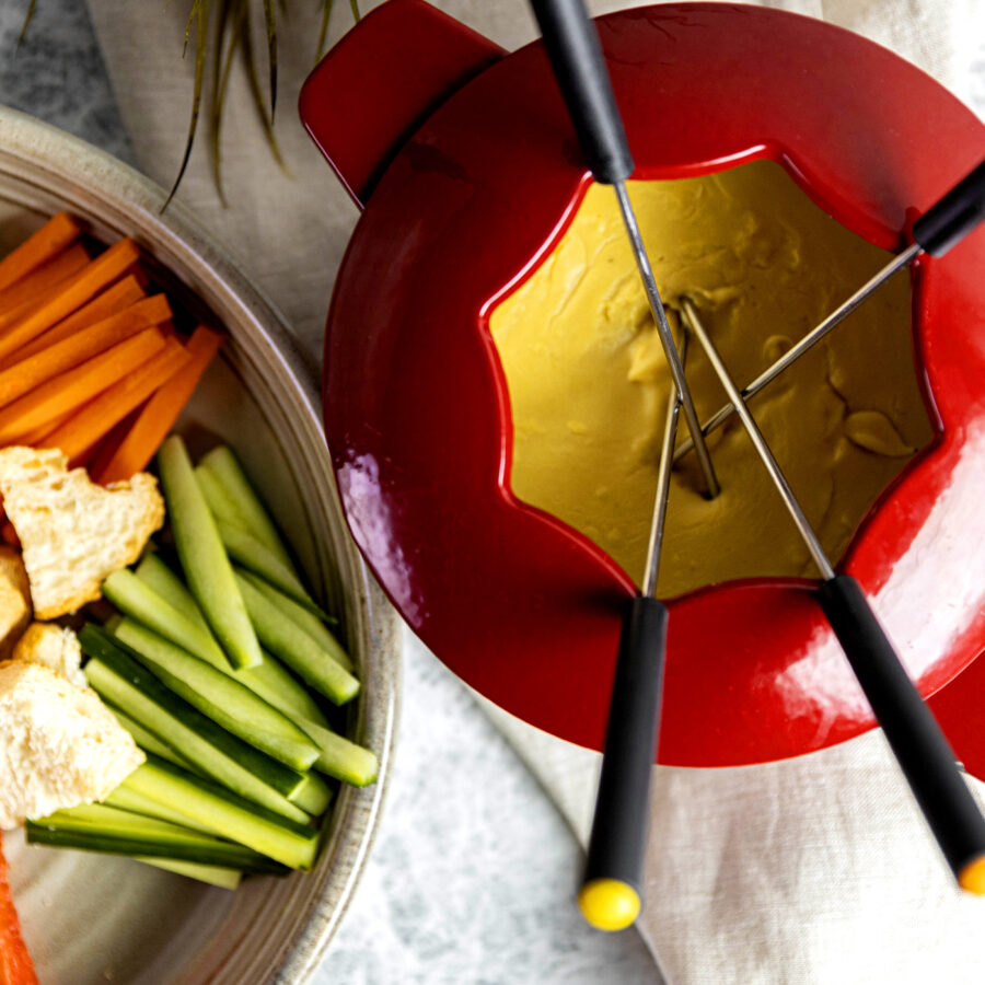 Red fondue pot with creamy plant-based cheese fondue and three forks, served with carrots, celery, and bread cubes on a light surface.