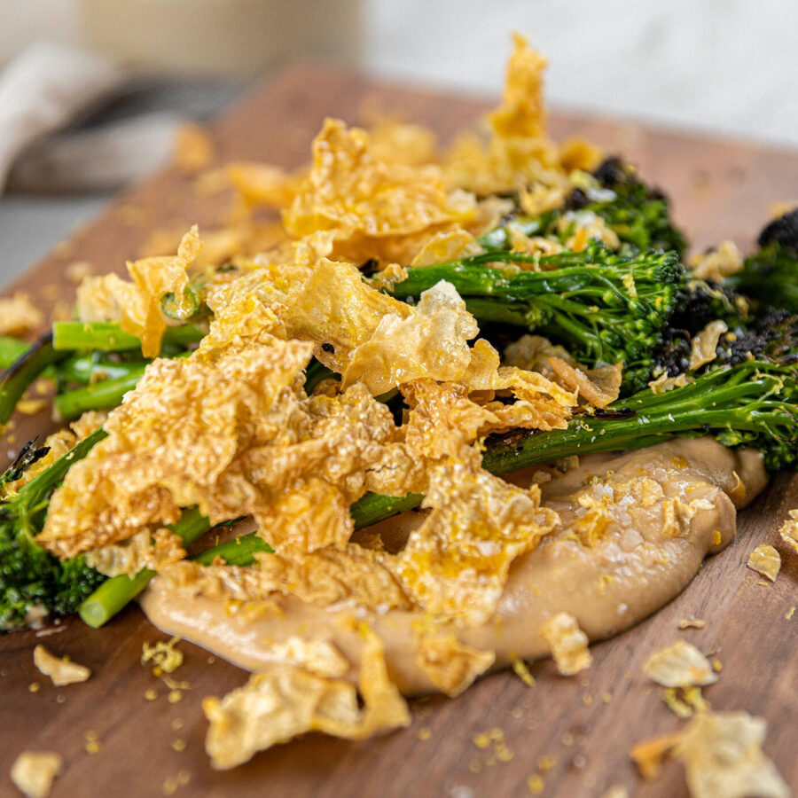 Broccolini on a wooden board, topped with plant-based crackling and served with a creamy hazelnut paste sauce.