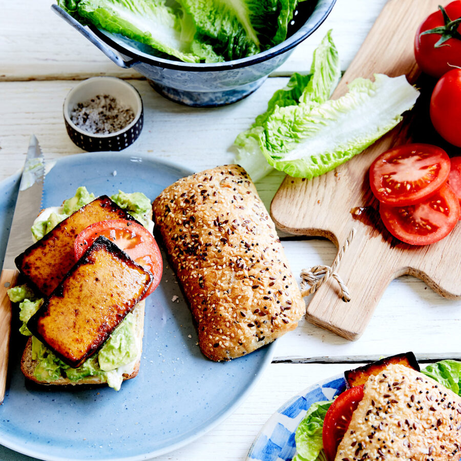 A seeded sandwich bap with grilled tofu and plant-based pork, lettuce, and tomato on a plate with fresh veg and another sandwich.