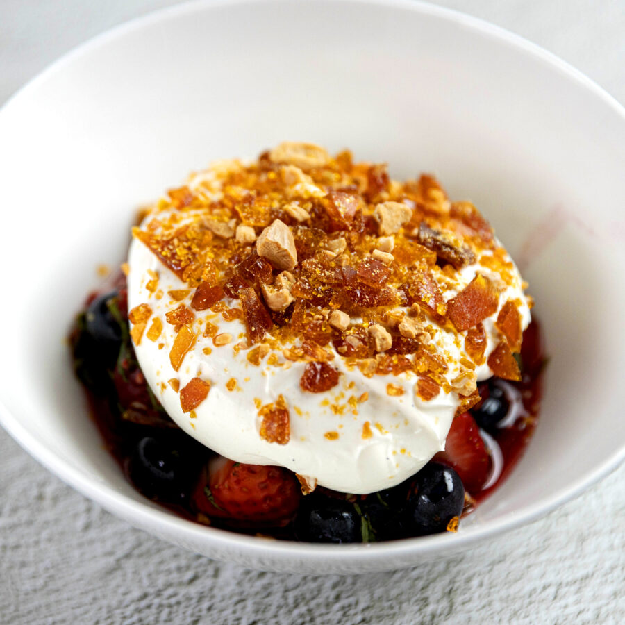 A white bowl filled with plant-based vanilla cream, topped with almond caramel praline over summer fruits like strawberries and blueberries.