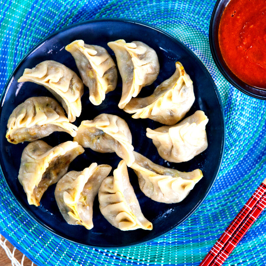 A plate of steamed plant-based cabbage momos arranged in a circle, served with red dipping sauce and red chopsticks beside the plate.