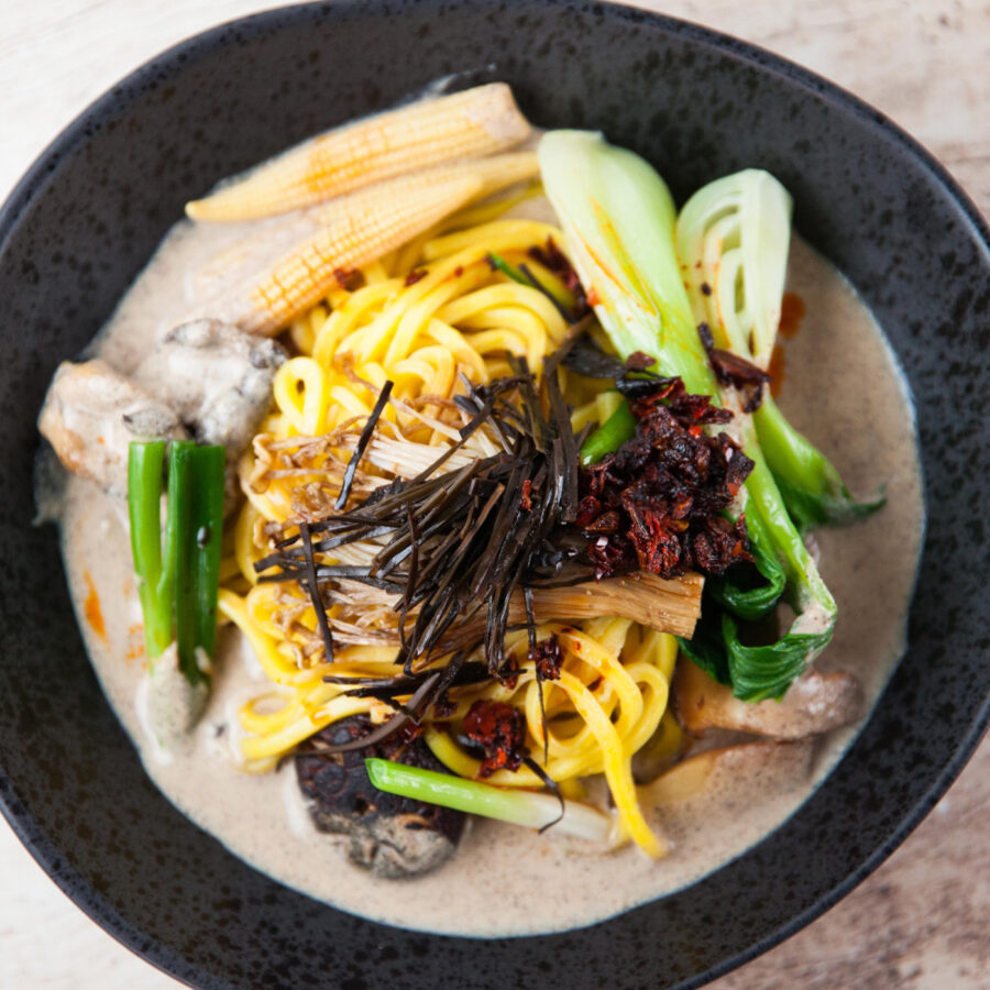 An overhead shot of ramen noodles in a bowl in a creamy broth, served with baby corn and Asian greens.