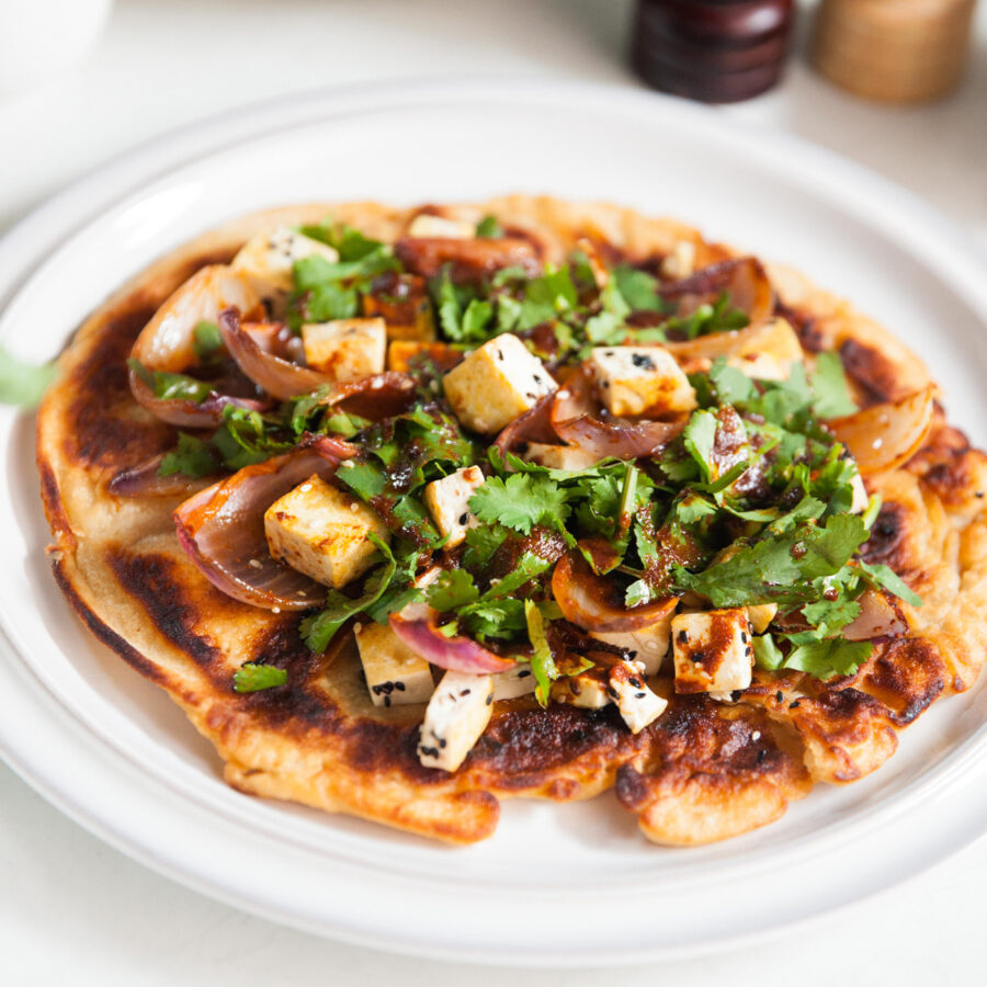 A cooked flatbread topped with cubes of plant-based paneer, spiced onions, fresh coriander, and spices on a white plate.