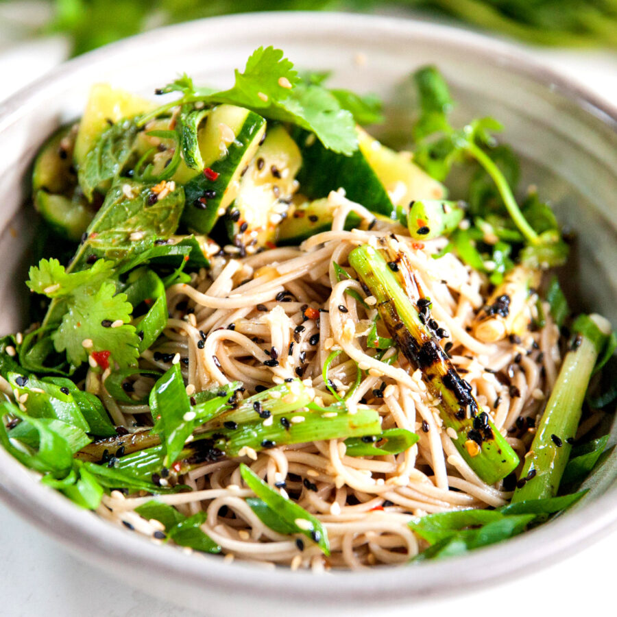 A bowl of soba noodles topped with grilled asparagus, crushed cucumber, coriander, spring onions, sesame seeds, and lime wedges.