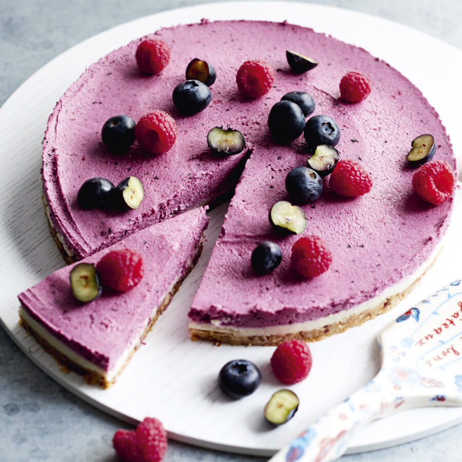 A round raw vegan blueberry cheesecake with a slice cut out, topped with fresh fruit, sits on a white serving board.
