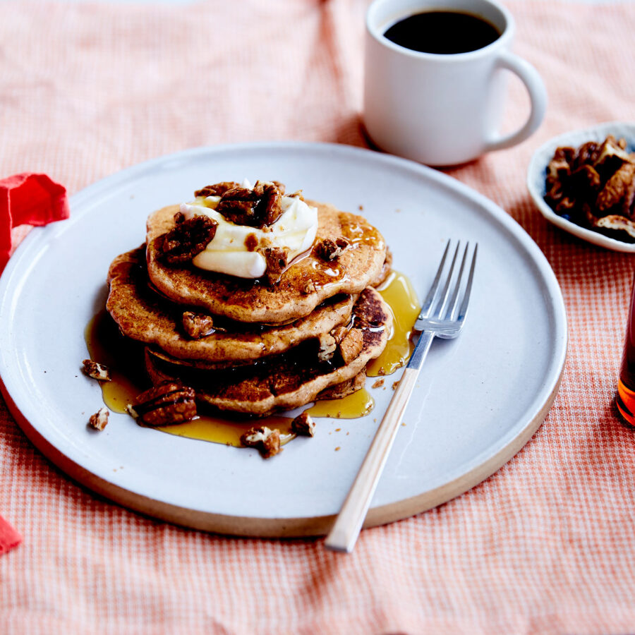 A stack of Spiced Pumpkin Pancakes topped with vegan butter and maple pecans, drizzled with syrup, sits on a plate with a fork. A cup of coffee, a small dish of pecans, and a syrup bottle are beside the plate.