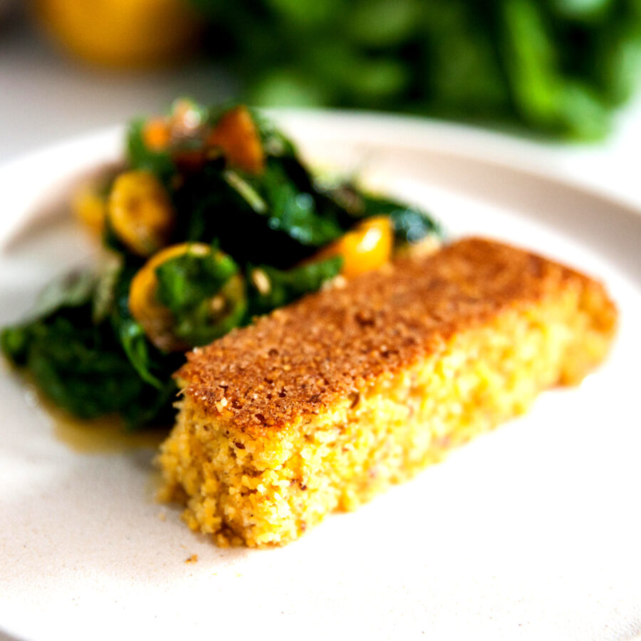 A slice of cornbread and a serving of leafy greens with yellow tomatoes and courgette on a white plate, all plant-based.
