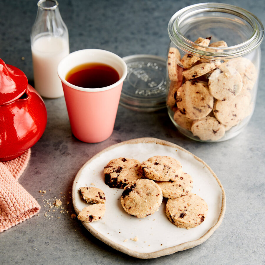 A plate of chocolate chip biscuits sits on a grey surface next to a jar of biscuits, a cup of tea or coffee, a bottle of plant-based milk, a red container, and a knitted cloth.