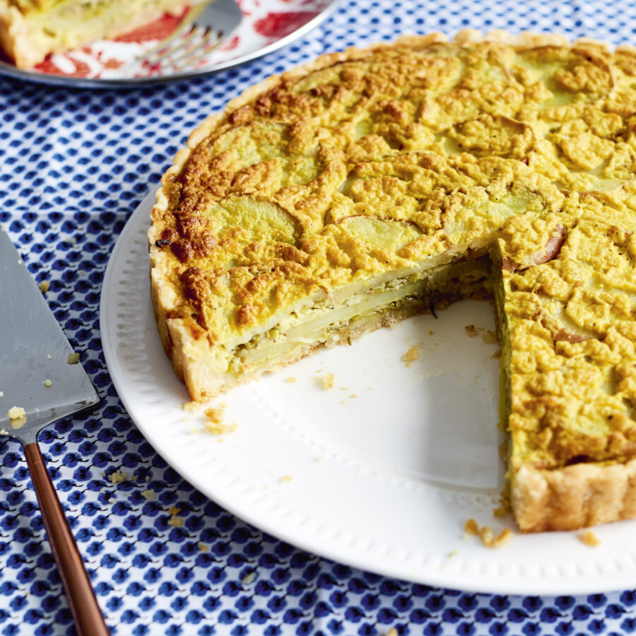 A sliced savoury plant-based leek and potato tart on a white plate with a serving utensil, set on a blue and white patterned tablecloth.