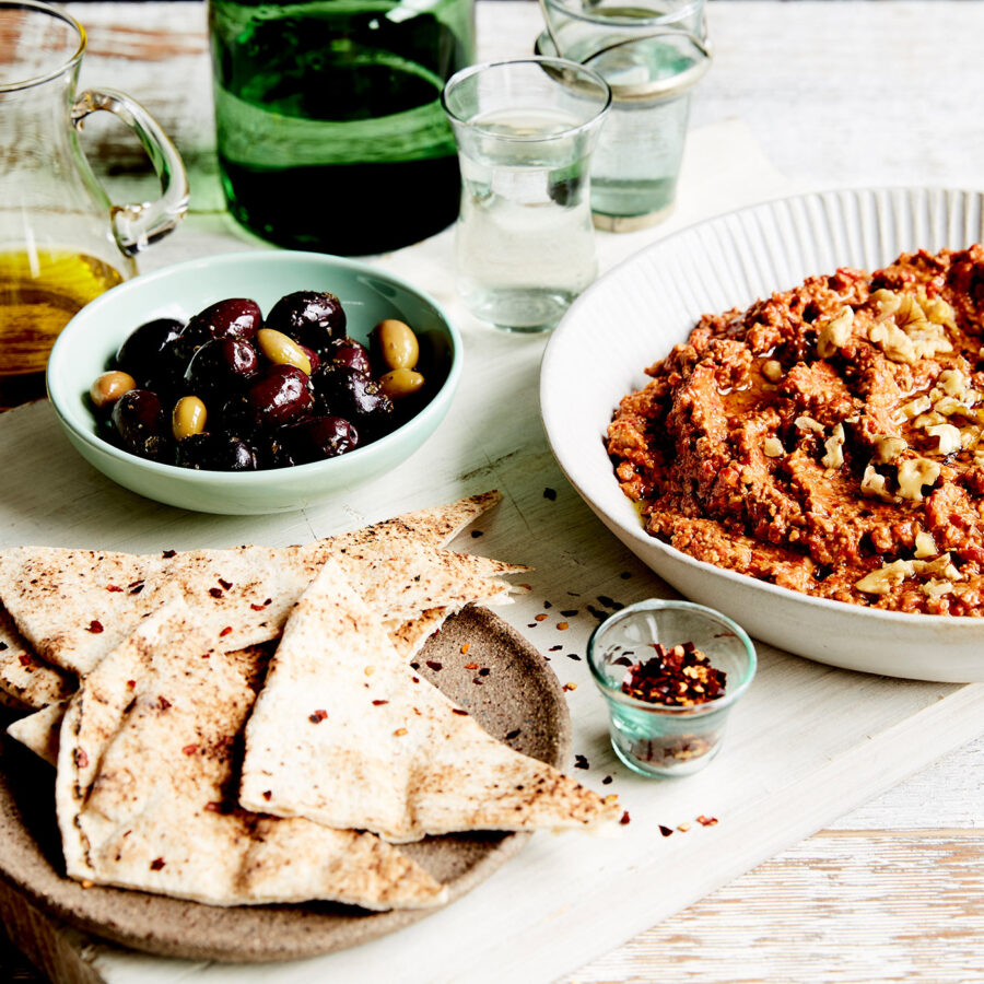 A chunky red dip sits to the left of a table setting, surrounded by pita bread and olives.