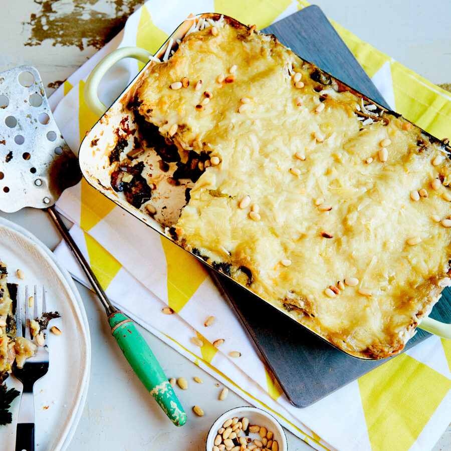 A baked mushroom lasagne in a rectangular dish, topped with vegan cheese, kale, and pine nuts, is partly served onto a plate; cutlery and scattered pine nuts are nearby.