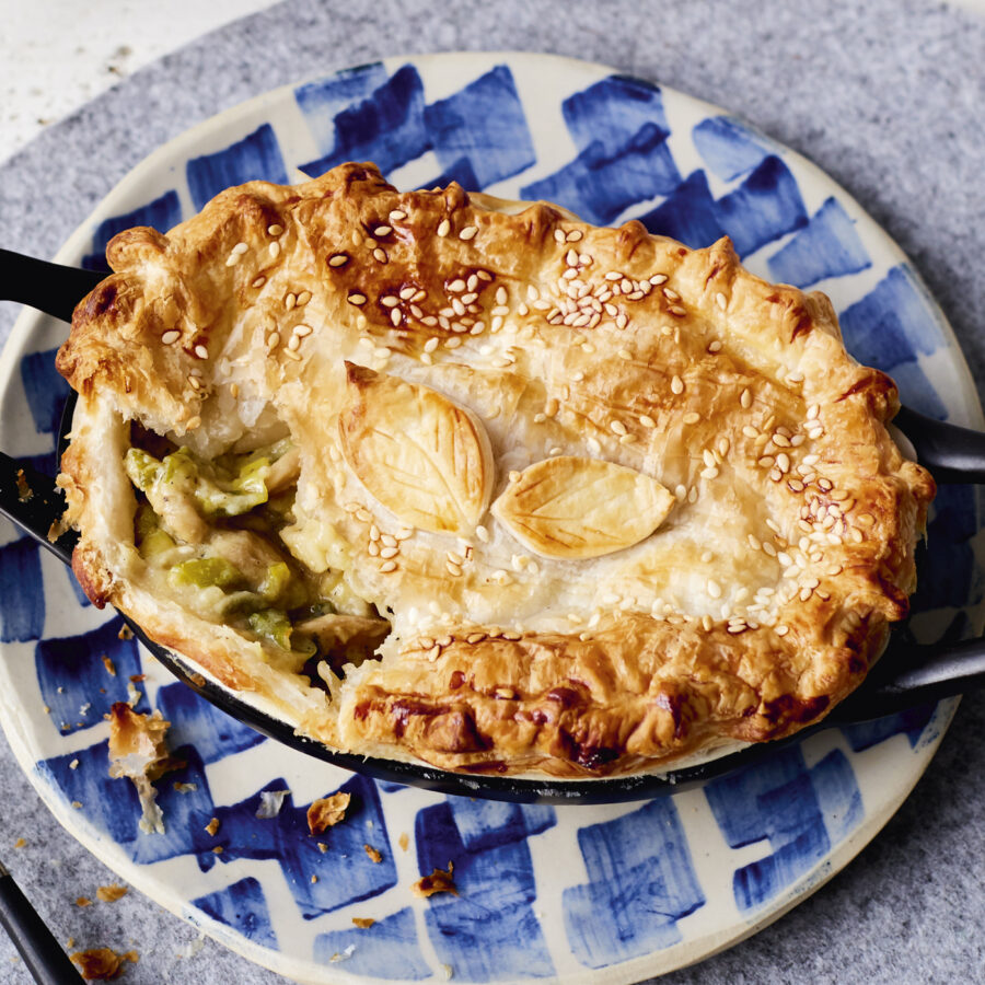 A golden-brown mushroom and leek pie with sesame seeds and leaf-shaped pastry sits on a plate, showing its plant-based filling inside.