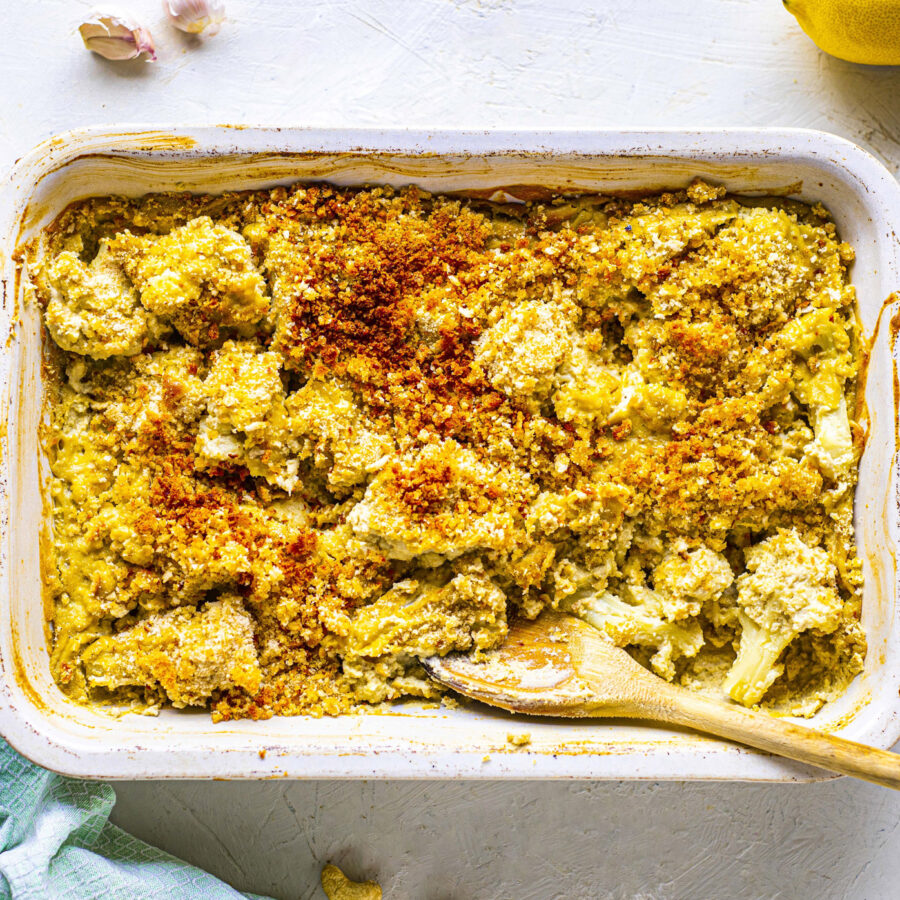 A rectangular oven tray filled with a very creamy looking cauliflower bake. It's topped with breadcrumbs. A wooden spoon sits in the corner.