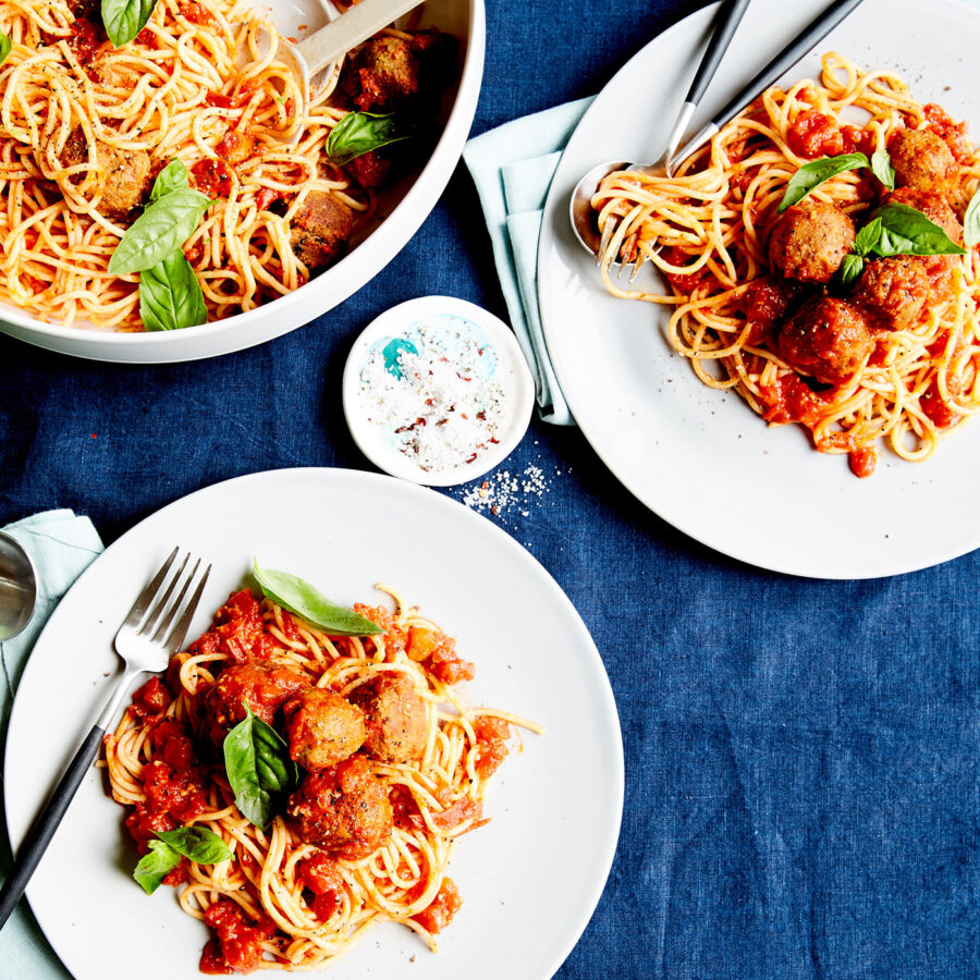 Plates of spaghetti with Napoli sauce and plant-based lentil almond meatballs, garnished with basil on a blue tablecloth.