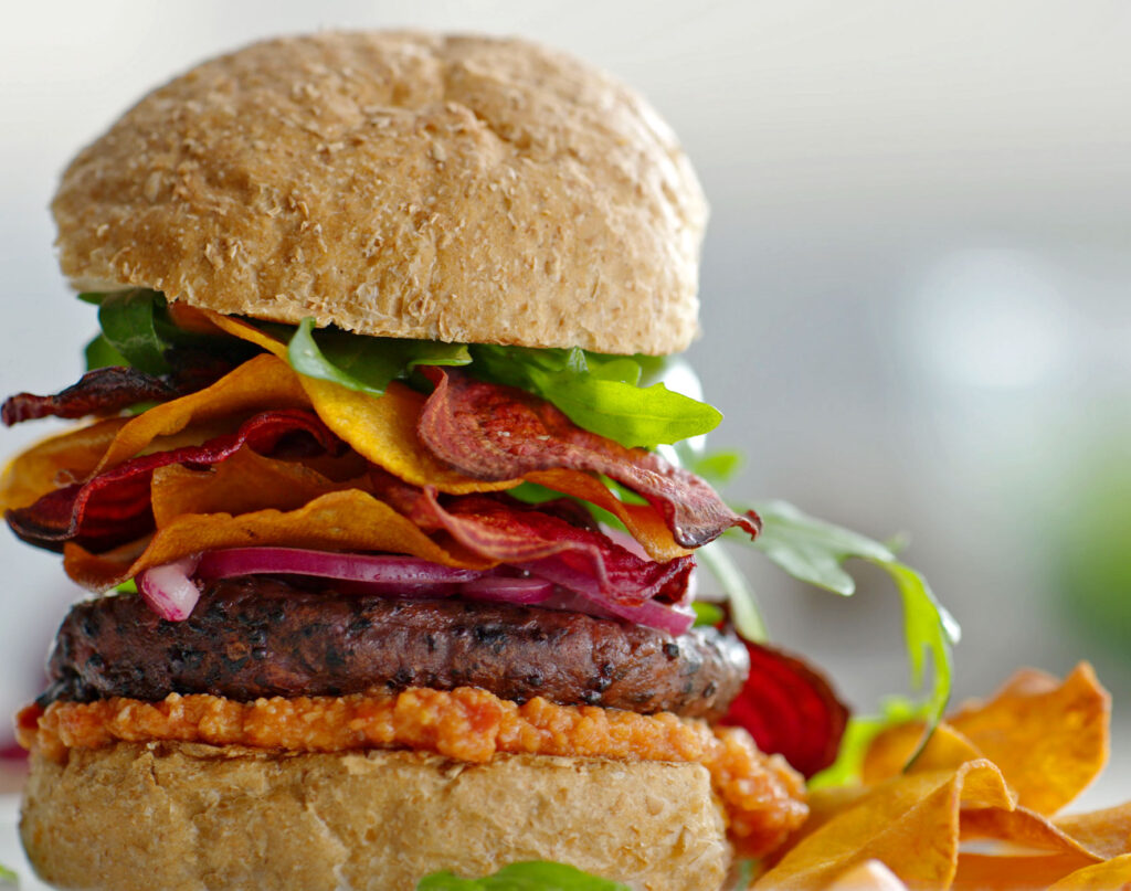 A close-up of a gourmet burger with a wholemeal bun, plant-based patty, leafy greens, red onions, vegetable crisps, and tomato sauce.