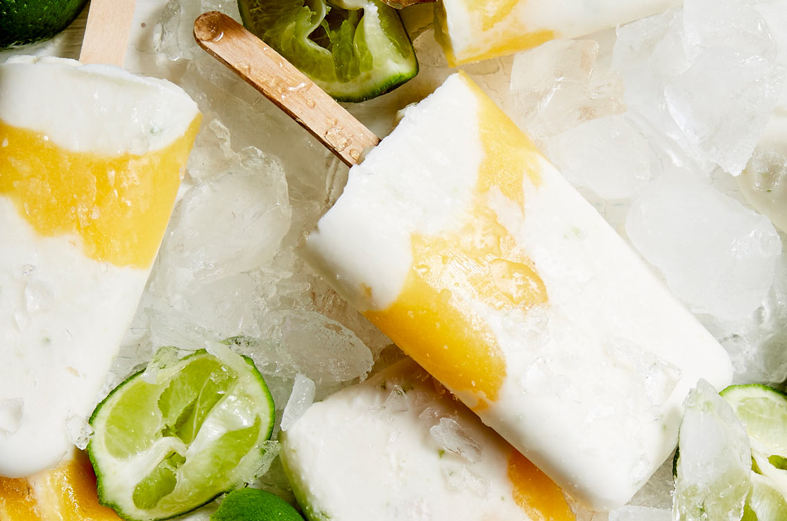 Plant-based creamy white and yellow ice lollies rest on ice, surrounded by sliced limes.