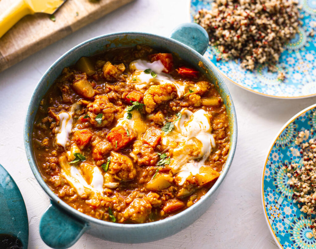 A bowl of veggie stew garnished with plant-based yoghurt and herbs, served alongside cooked quinoa on a patterned plate.
