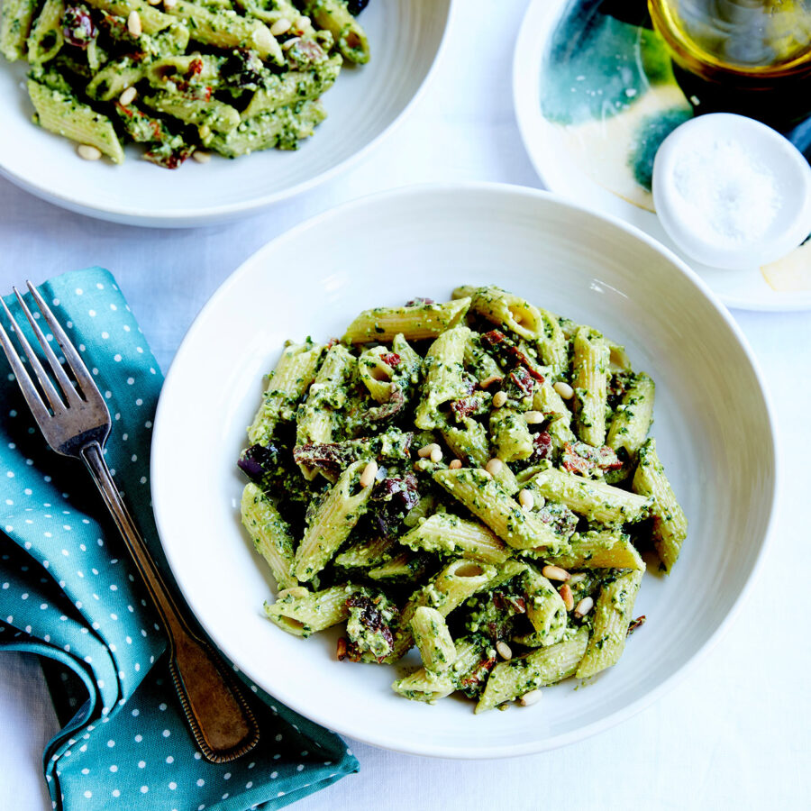A white bowl of vegan pesto pasta with sundried tomatoes, pine nuts, and olives sits on a white table next to a fork and teal serviette.