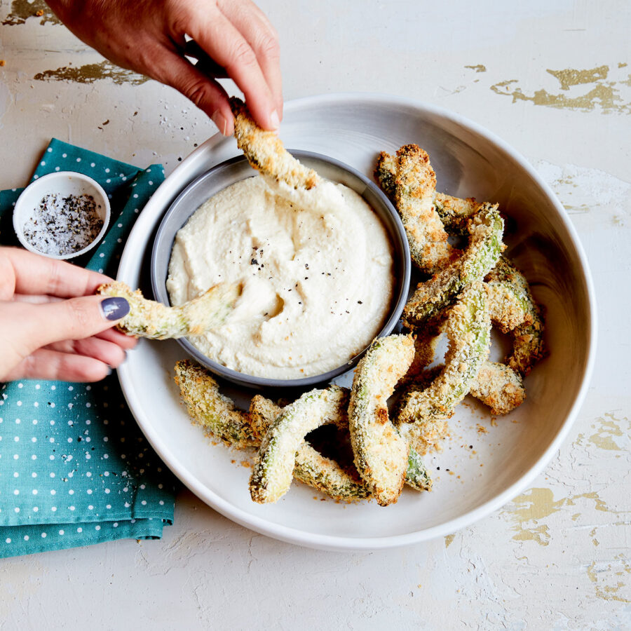 Hands dipping crunchy Avocado Crisps into a bowl of plant-based creamy dip, with more crisps around it, next to a blue serviette and seasoning.
