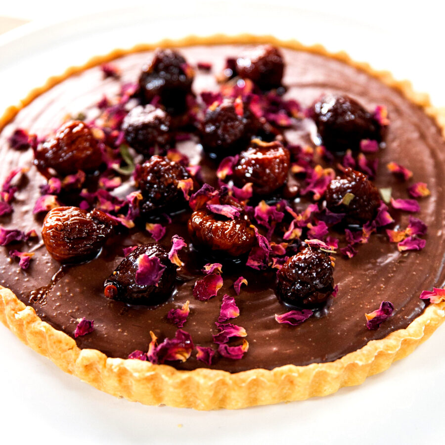 A plant-based chocolate tart topped with cherries and dried rose petals on a white plate—a perfect choice for romantic recipes.
