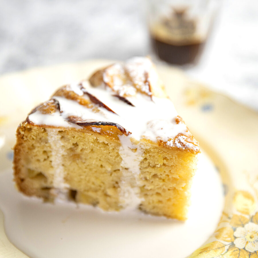A slice of glazed lemon olive oil cake with vegan white icing rests on a floral-patterned plate, a glass of dark liquid behind.