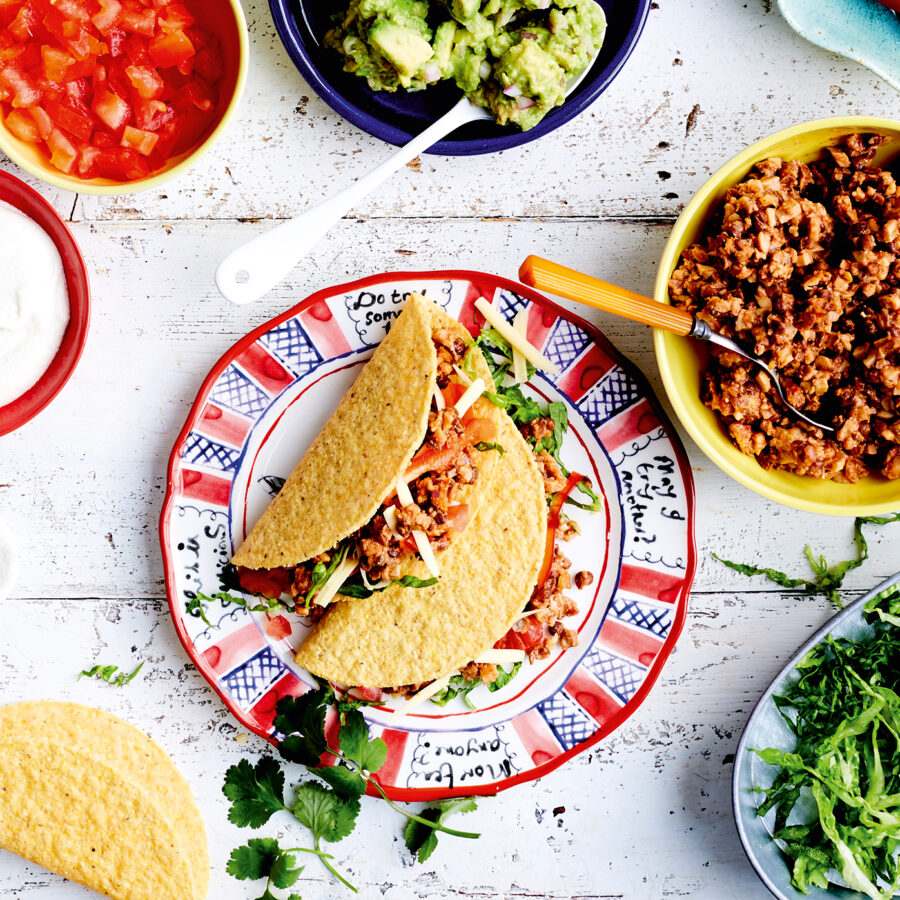A table set with two vegetarian tacos filled with plant-based mince, shredded lettuce, and vegan cheese, surrounded by salsa, guacamole, dairy-free soured cream, and extra taco accompaniments.