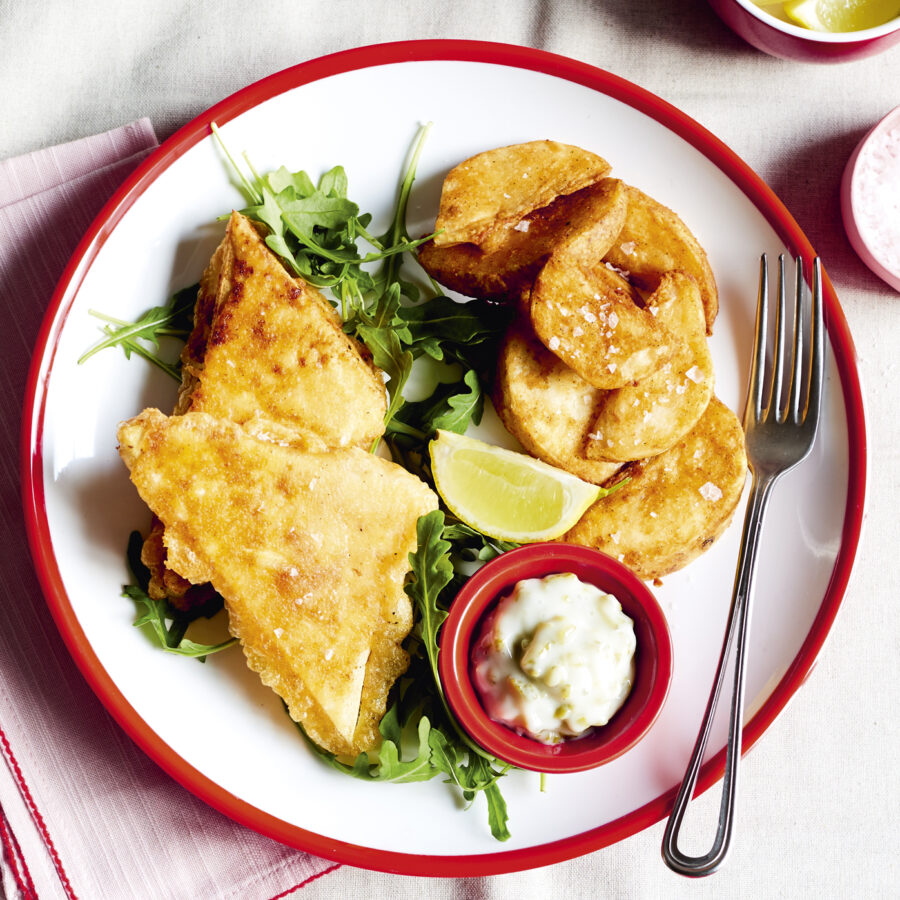 A plate with breaded vegan fish-style fillets on rocket, potato wedges, a lemon wedge, tartare sauce, tofu pieces, and a fork.