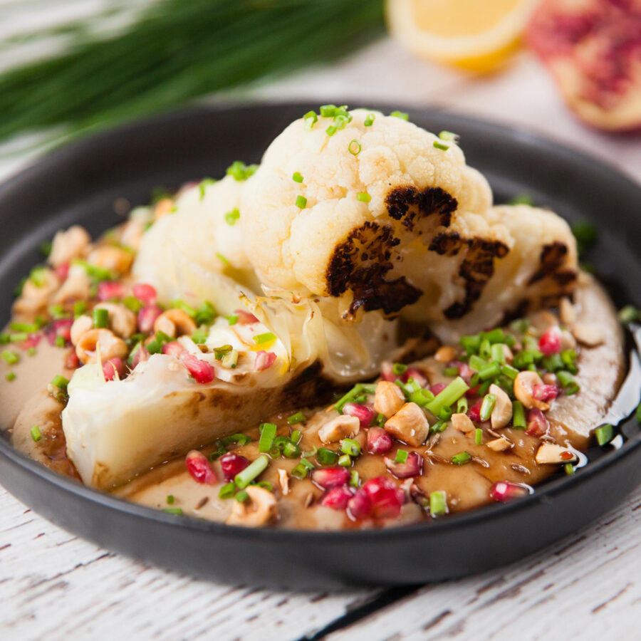 A cauliflower wedge topped with chopped nuts, pomegranate seeds, fresh herbs, and pomegranate molasses sits on a rustic white table.