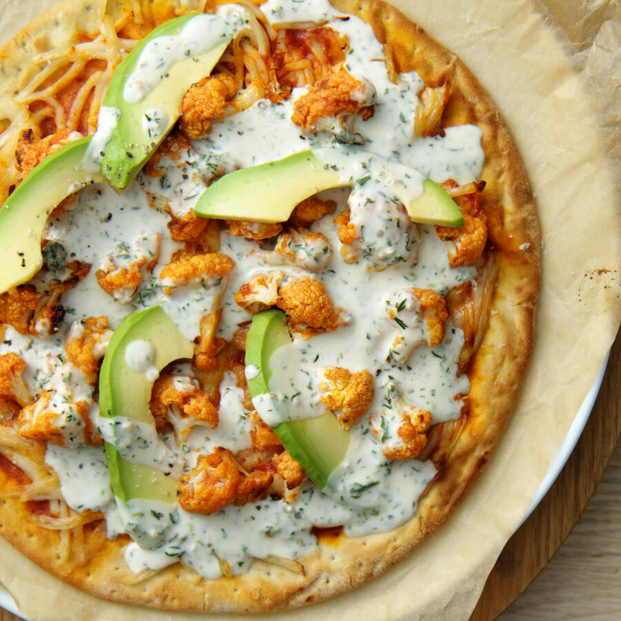 Buffalo cauliflower pizza topped with avocado slices, seasoned cauliflower, and plant-based creamy herb sauce on baking parchment.