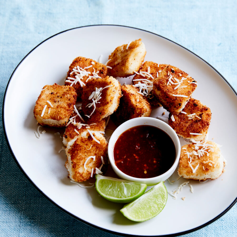 A plate with coconut-crusted tofu cubes, garnished with grated vegan cheese, served with chilli lime sauce and lime wedges.