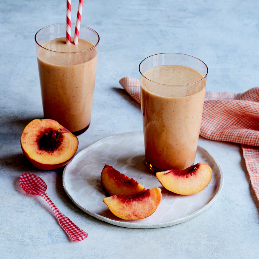 Two glasses of plant-based Peaches & Cream Smoothie with red striped straws, peach slices, and a pink spoon on a white plate, orange cloth behind.