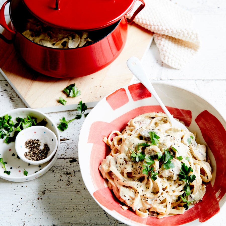 A bowl of creamy plant-based Mushroom Fettuccine Alfredo with parsley sits on a white table beside a red pot, herbs, and pepper.