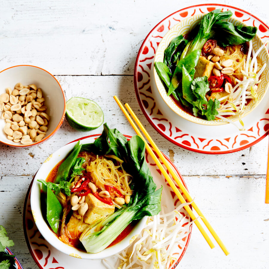 Two bowls of Malaysian-inspired vegan laksa with tofu, pak choi, bean sprouts, peanuts, and herbs on a white table with lime.