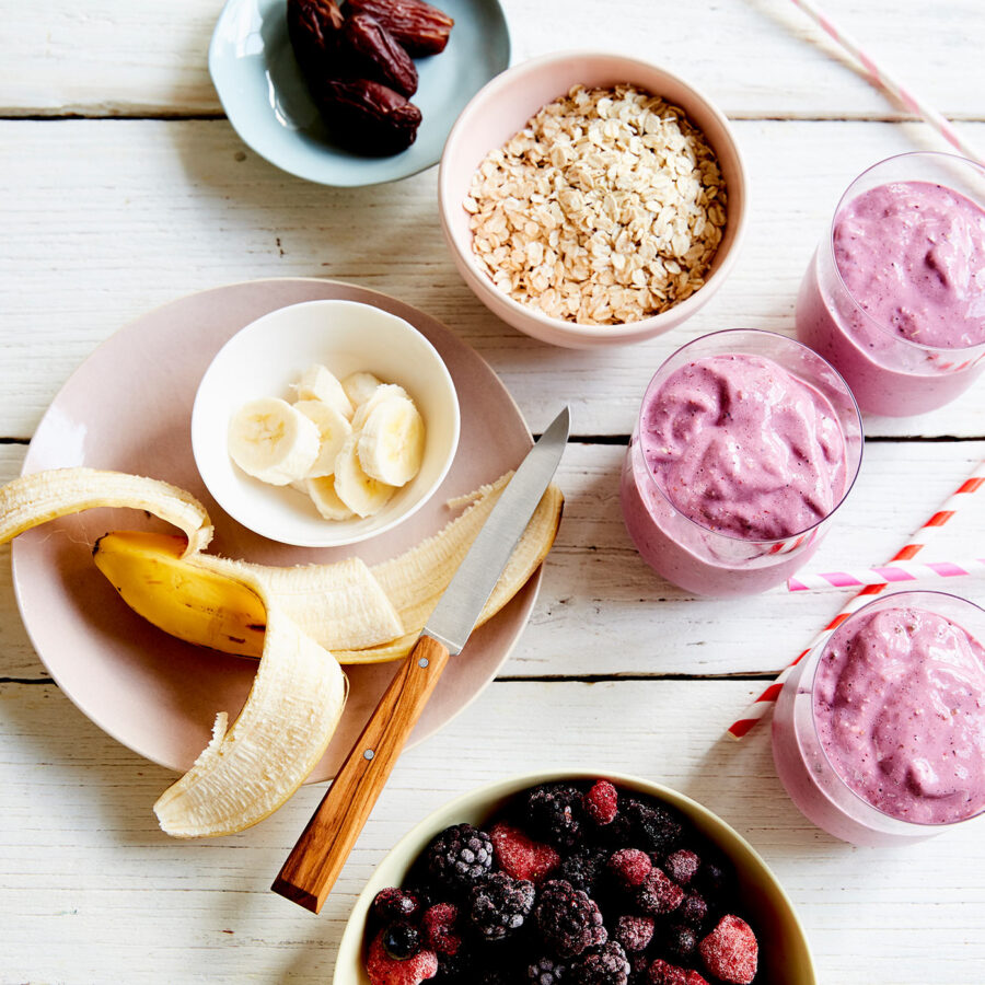 A banana, sliced banana, oats, dates, mixed berries, and three glasses of plant-based breakfast smoothie on a white table.