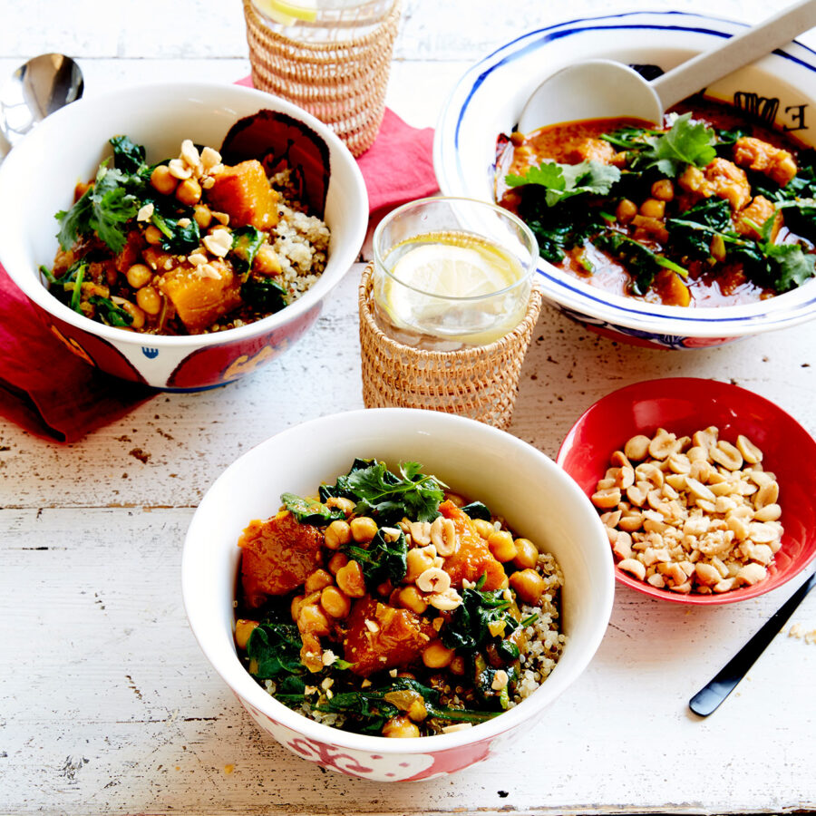 Three bowls of chickpea curry stew with greens, quinoa, chopped peanuts, lemon water, and cutlery on a white wooden table.