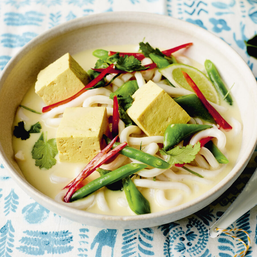 A bowl of Thai Coconut Soup with tofu cubes, mangetout, red pepper strips, coriander, and a light broth on a patterned tablecloth.