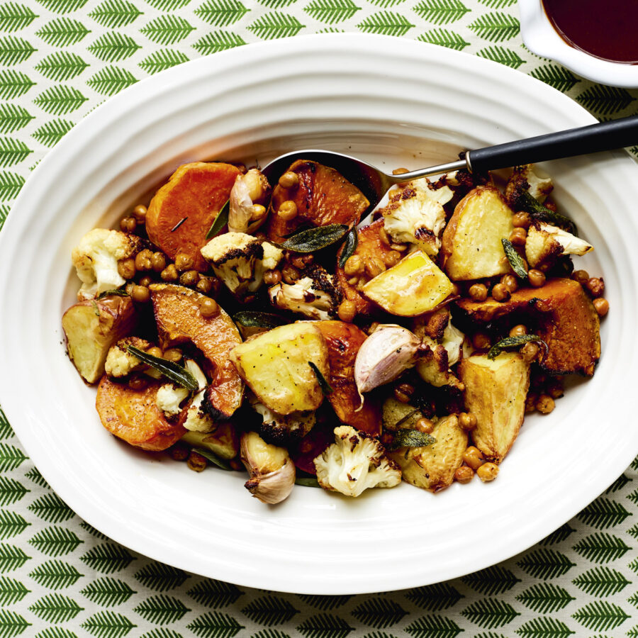 An overhead image of a bowl of herbed chickpeas sprinkled over roast potato and cauliflower. A 70s style patterned green and white tablecloth sits underneath.