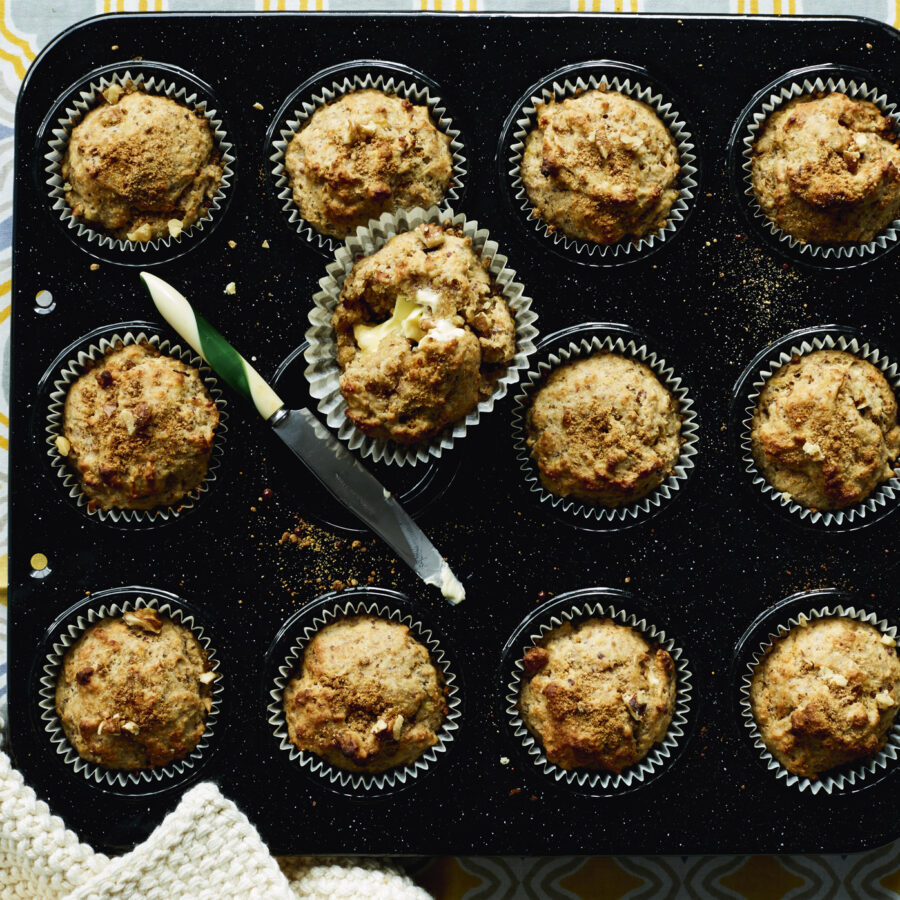 A muffin tin with twelve muffins, one partially unwrapped and spread with plant-based margarine; a butter knife rests beside it.