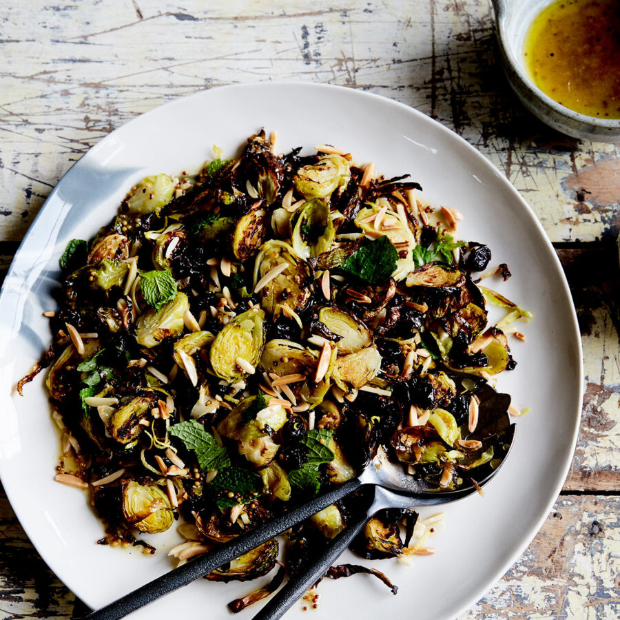 A plate of roasted Brussels sprouts with flaked almonds and mint on a wooden table, with plant-based dressing and mint nearby.
