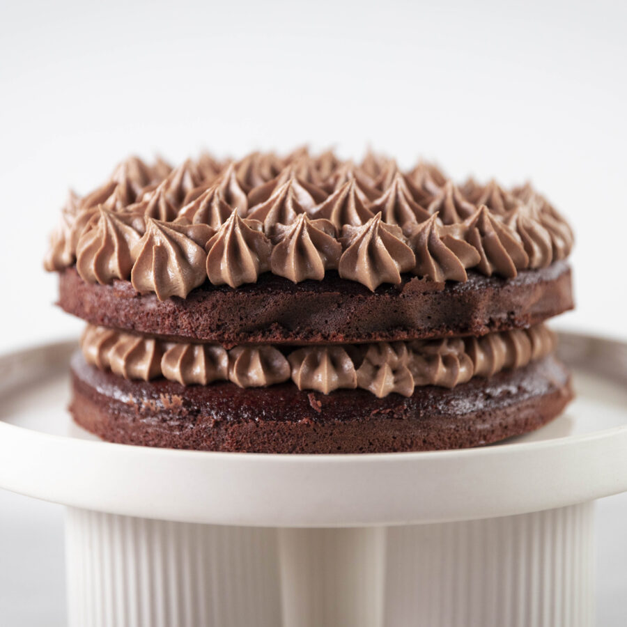 A decadent plant-based Chocolate Celebration Cake with two chocolate layers and vegan icing, elegantly displayed on a white cake stand.
