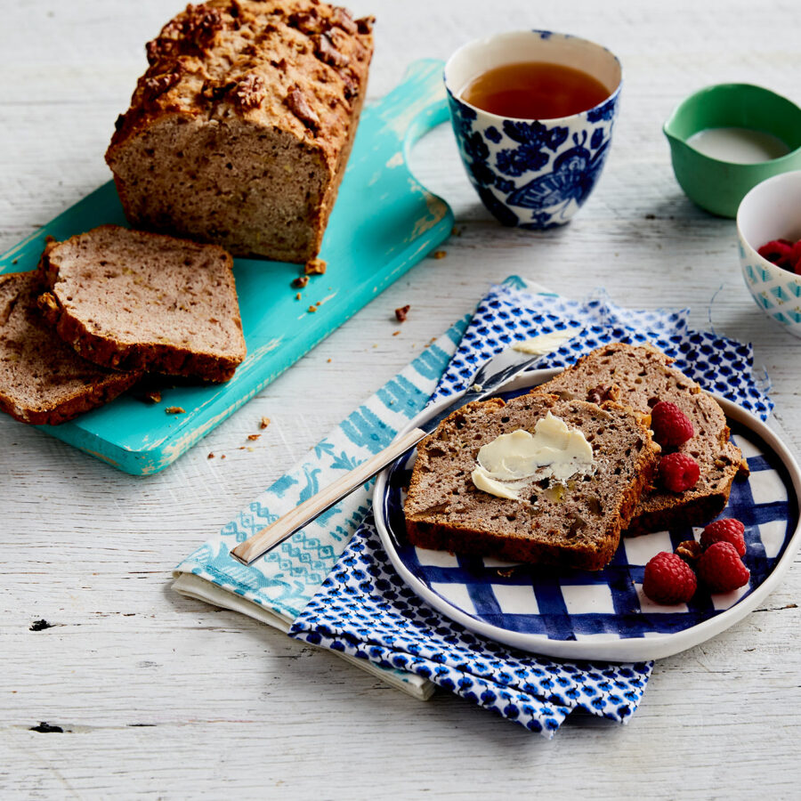 A plate with slices of Ultimate Banana Bread, one topped with plant-based butter and raspberries, sits beside tea and vegan milk.