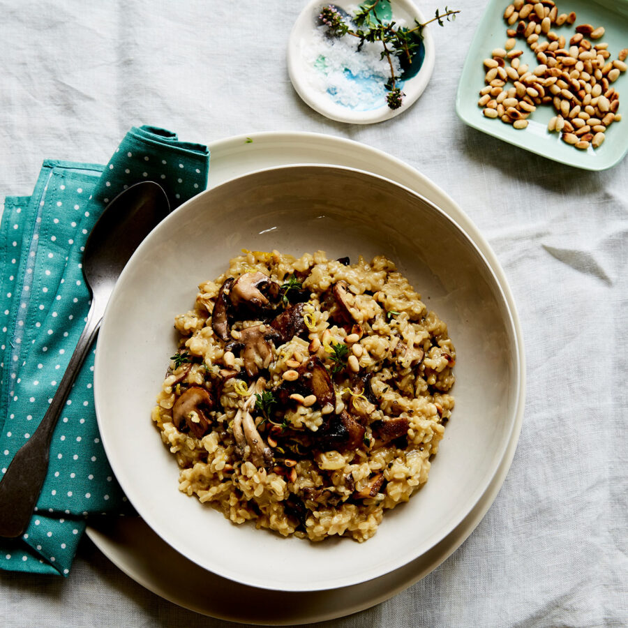 A bowl of creamy plant-based mushroom risotto on a white tablecloth, with pine nuts, teal serviette, spoon, and salt dish nearby.