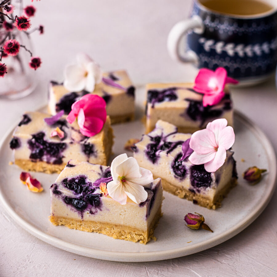A plate with five slices of plant-based baked cheesecake, each topped with edible flowers, sits by tea and pink blooms—a blueberry pudding.