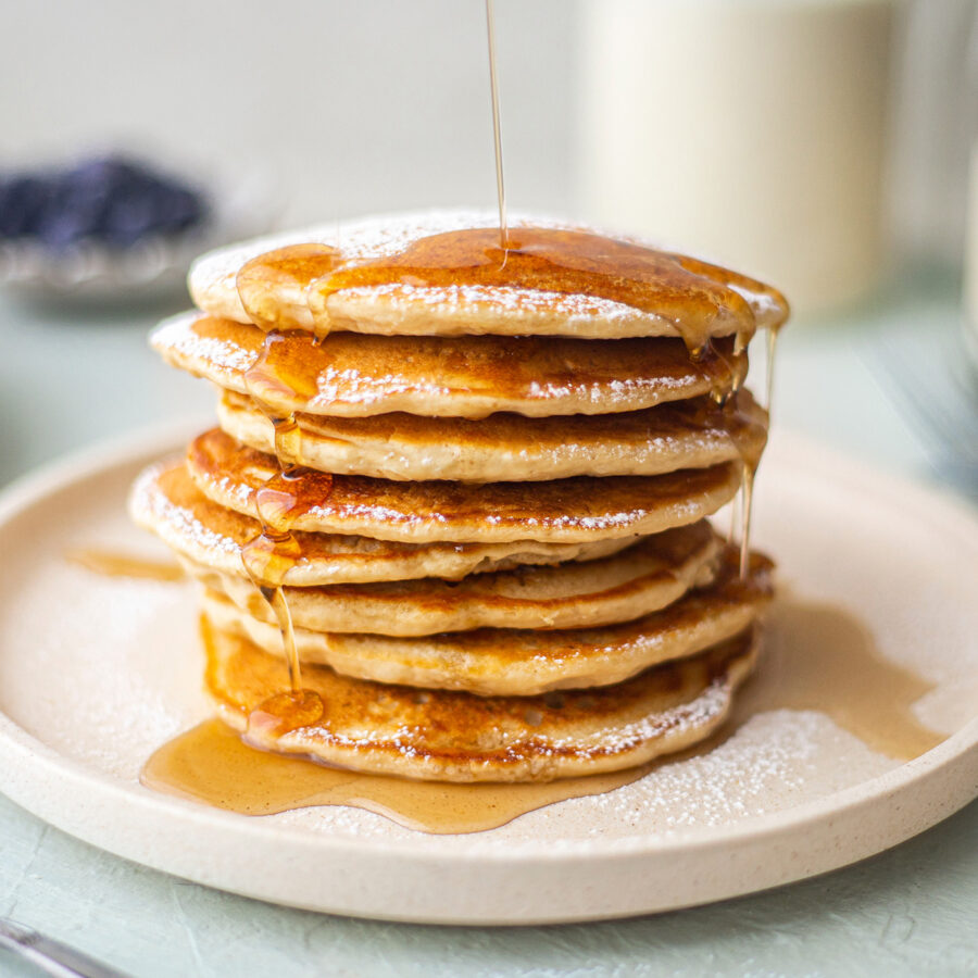 A stack of plant-based protein pancakes on a plate is topped with icing sugar, banana slices, and syrup being poured over them.