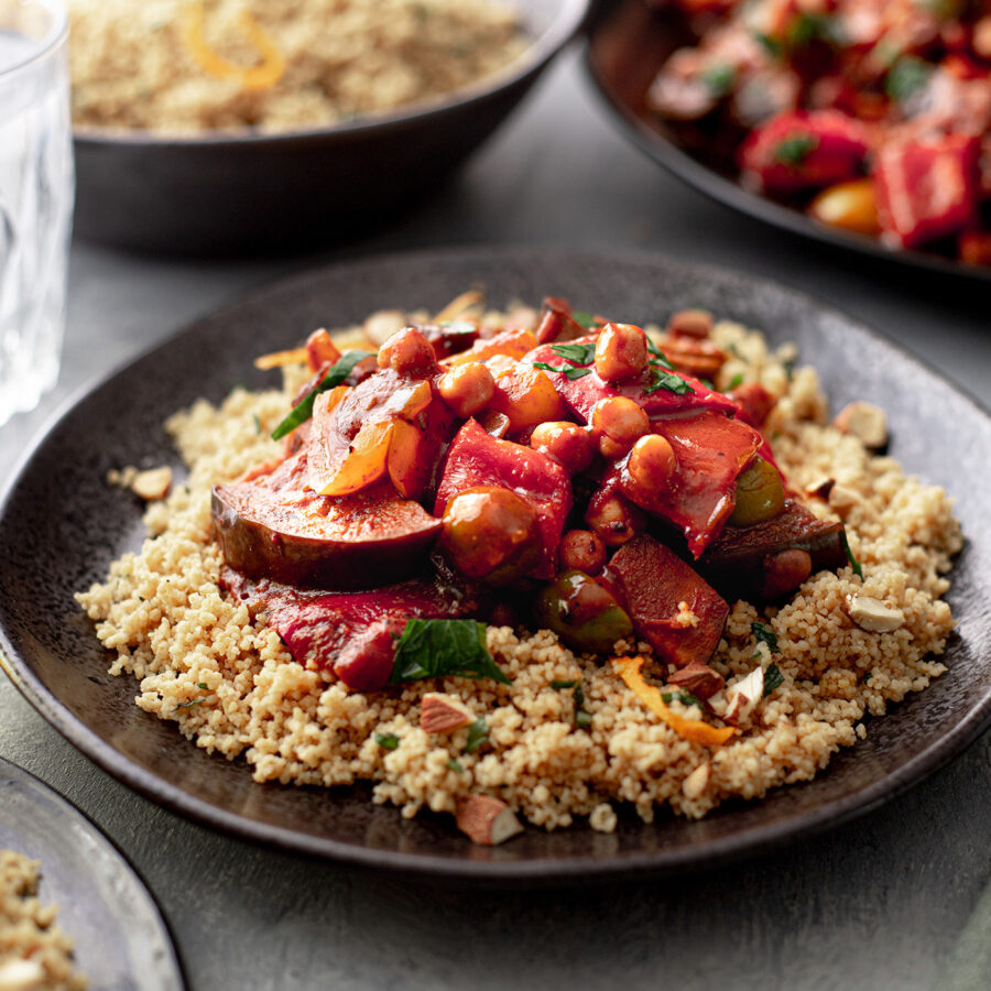 A plate of herbed couscous with chickpea and vegetable tagine, garnished with fresh herbs, on a dark plate and glass of water.