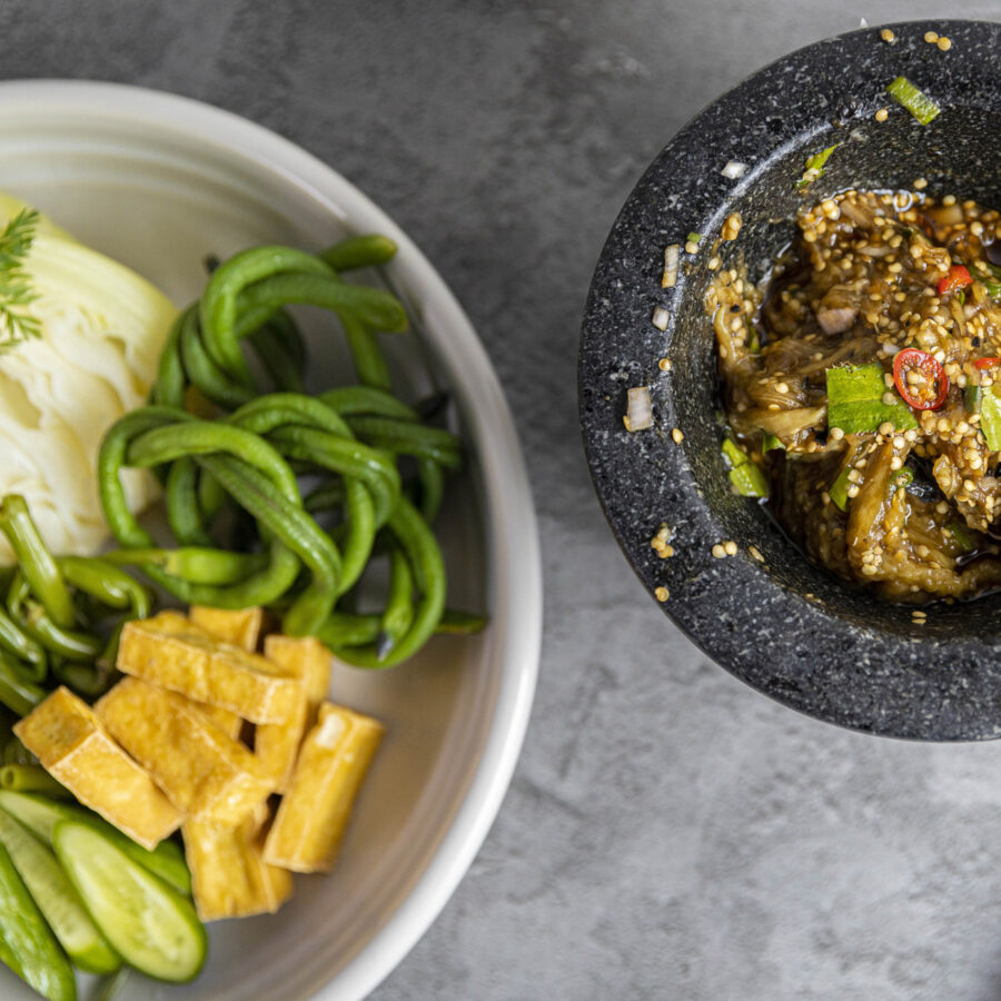 A plate of fresh vegetables and tofu slices sits beside a stone mortar filled with chunky Smoky Aubergine Relish.