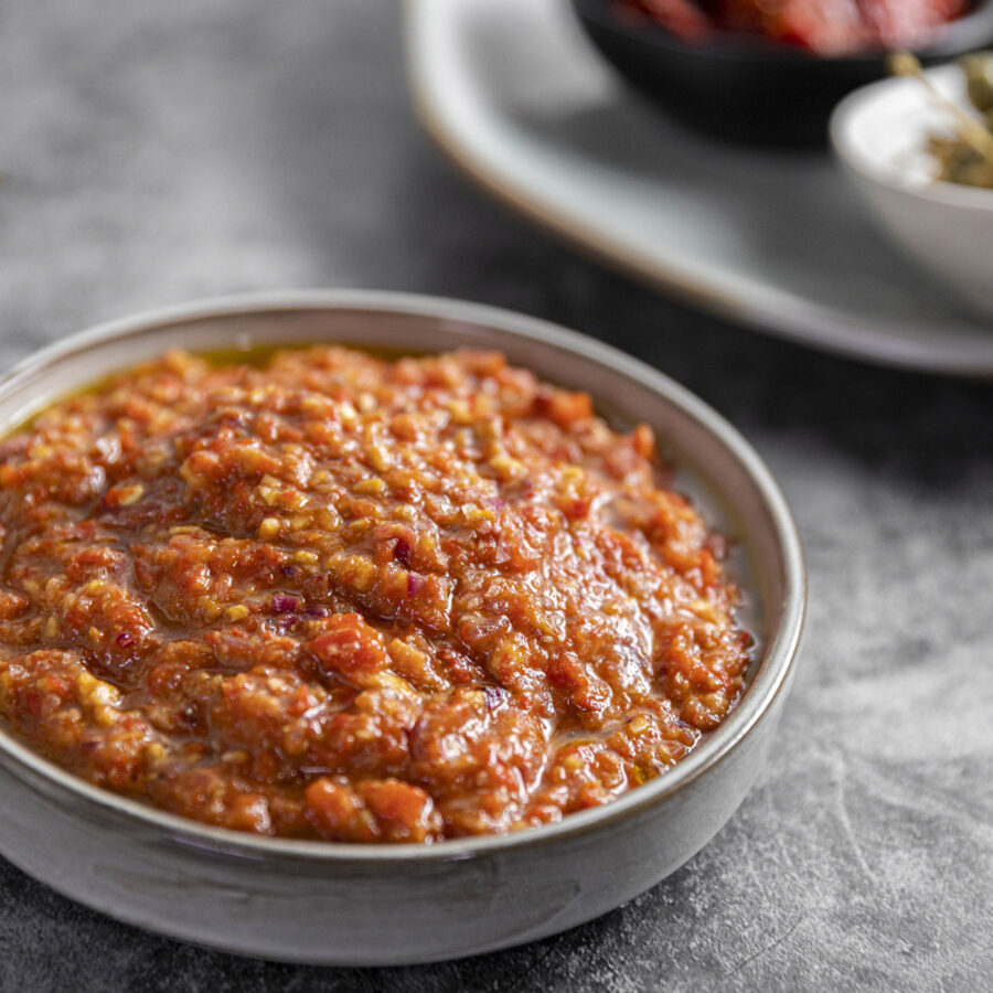 A shallow bowl filled with chunky plant-based chilli sauce sits on a grey surface, with blurred dishes in the background.